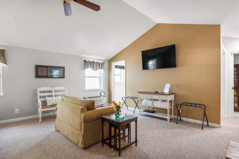 Suite Divine sitting area. Sitting area with flat screen TV mounted on the wall. Underneath is a farm style cabinet with a lamp and DVD player. A luggage rack it on the left. In the foreground are two comfortable upholstered chairs with a small table in between.