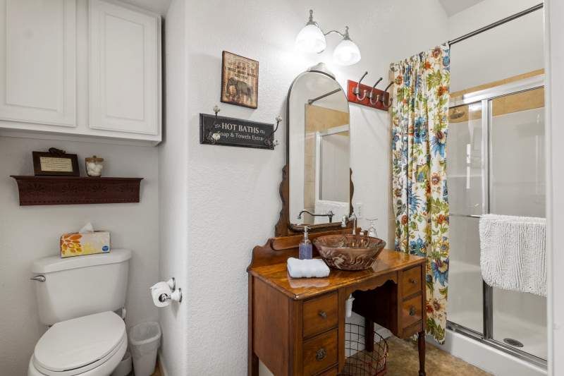Suite Divine Bathroom Bright lighting illuminates an antique dresser repurposed into a bathroom vanity with a glass surface mount sink. There is a step-in shower and white bath mat and hand towel.